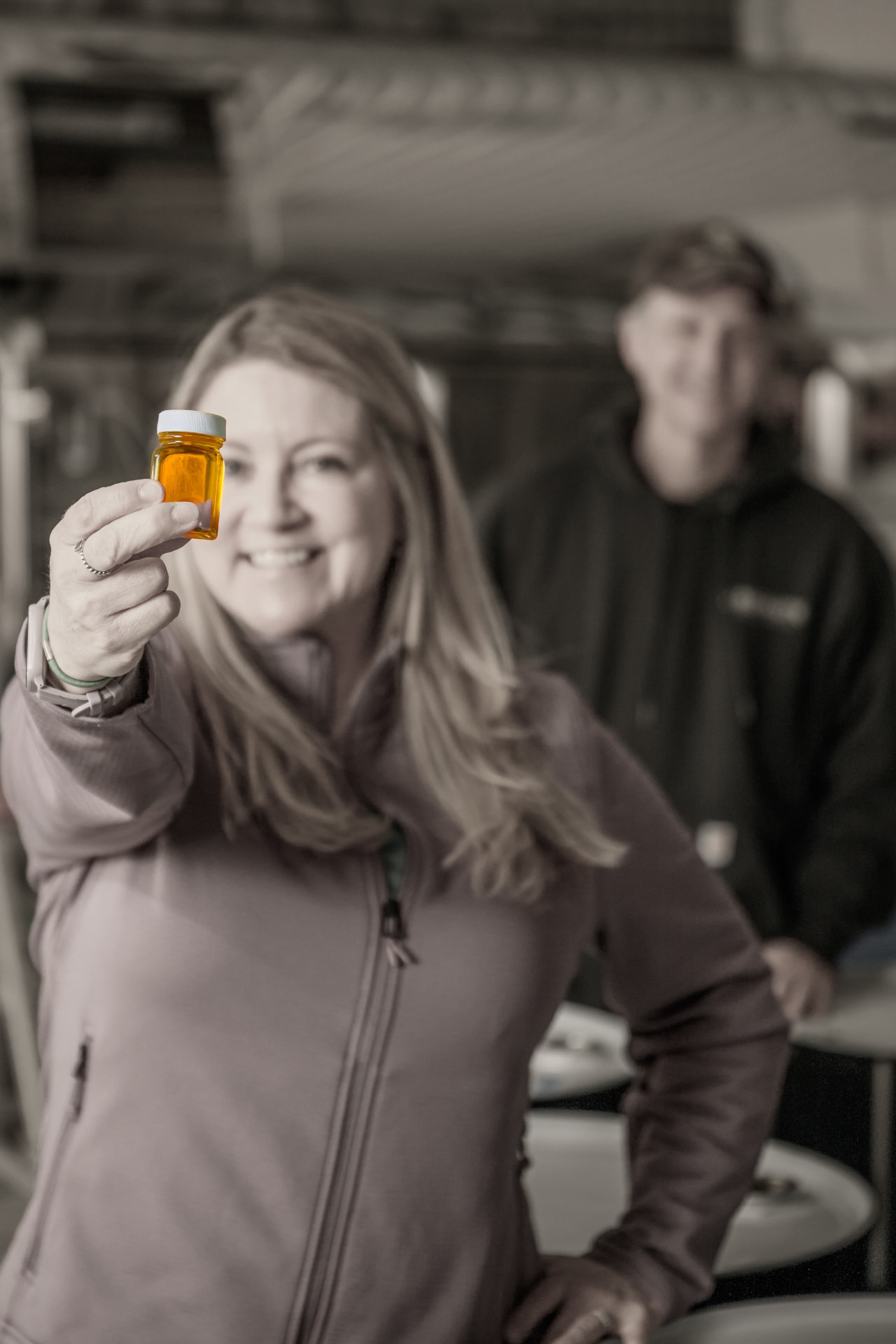 Lane holding up a syrup grade sample jar with Greg in the sugarhouse behind her